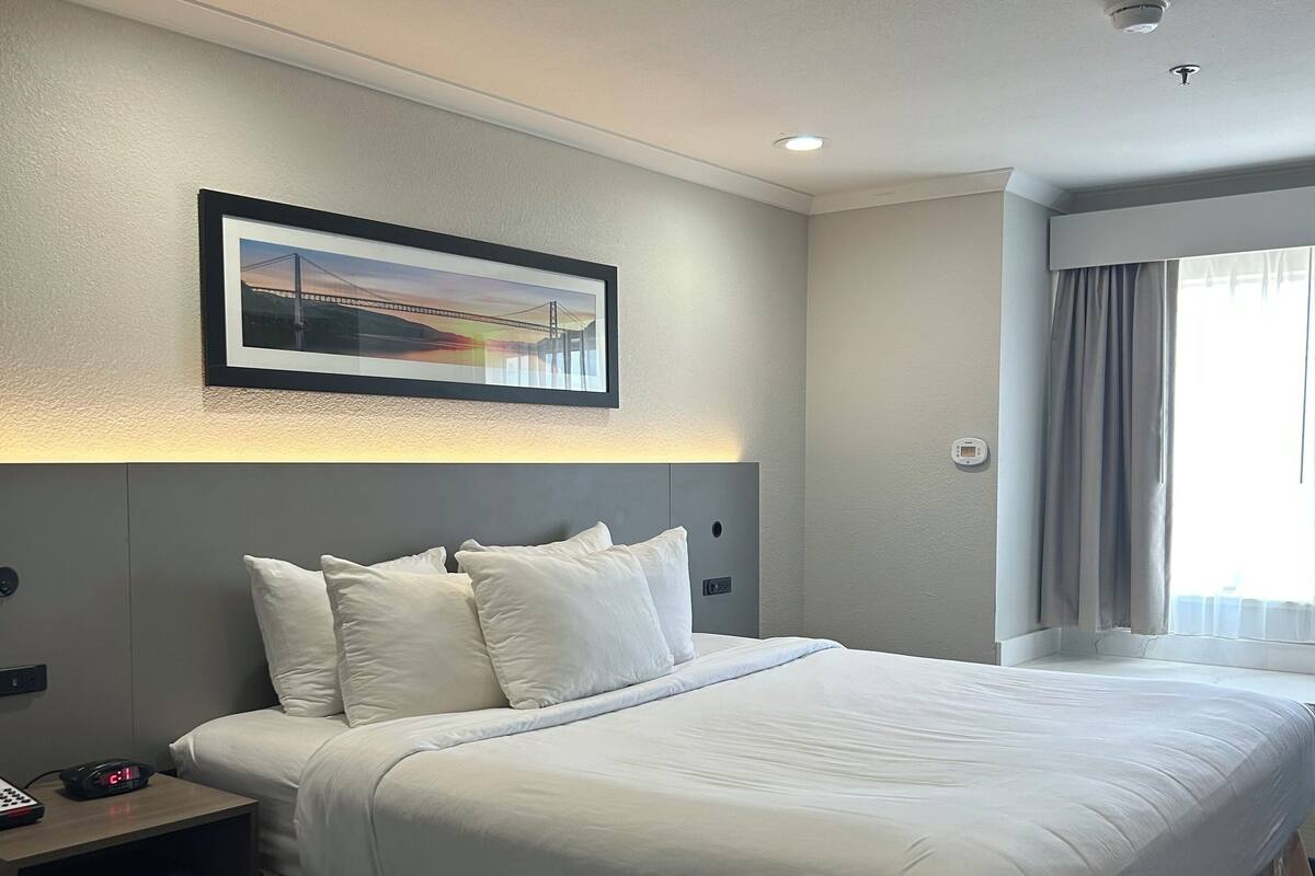 A hotel room with a large bed, a bedside table with a clock, a framed picture on the wall, and a window with curtains.