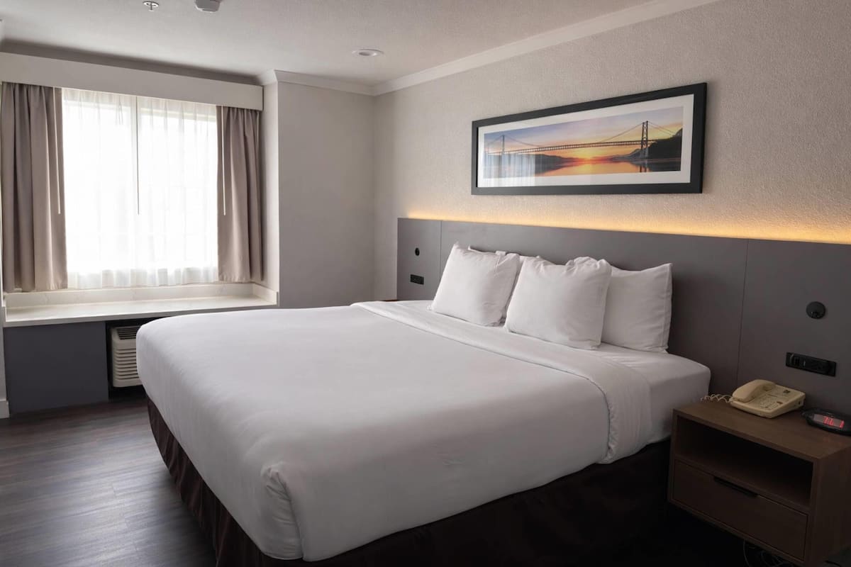 A hotel room with a large bed, a bedside telephone, a window with curtains, and a framed picture on the wall.