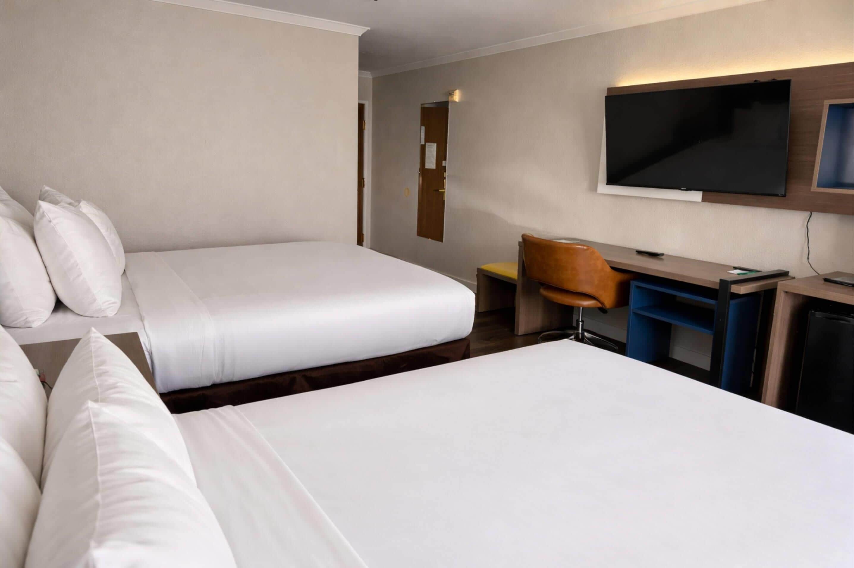 Quarto de hotel com duas camas, uma escrivaninha, uma cadeira, uma televisão e uma porta.