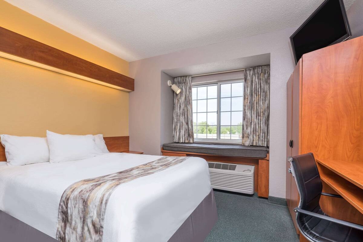A hotel room with a bed, a desk, a chair, a window with curtains, and a television on a stand.