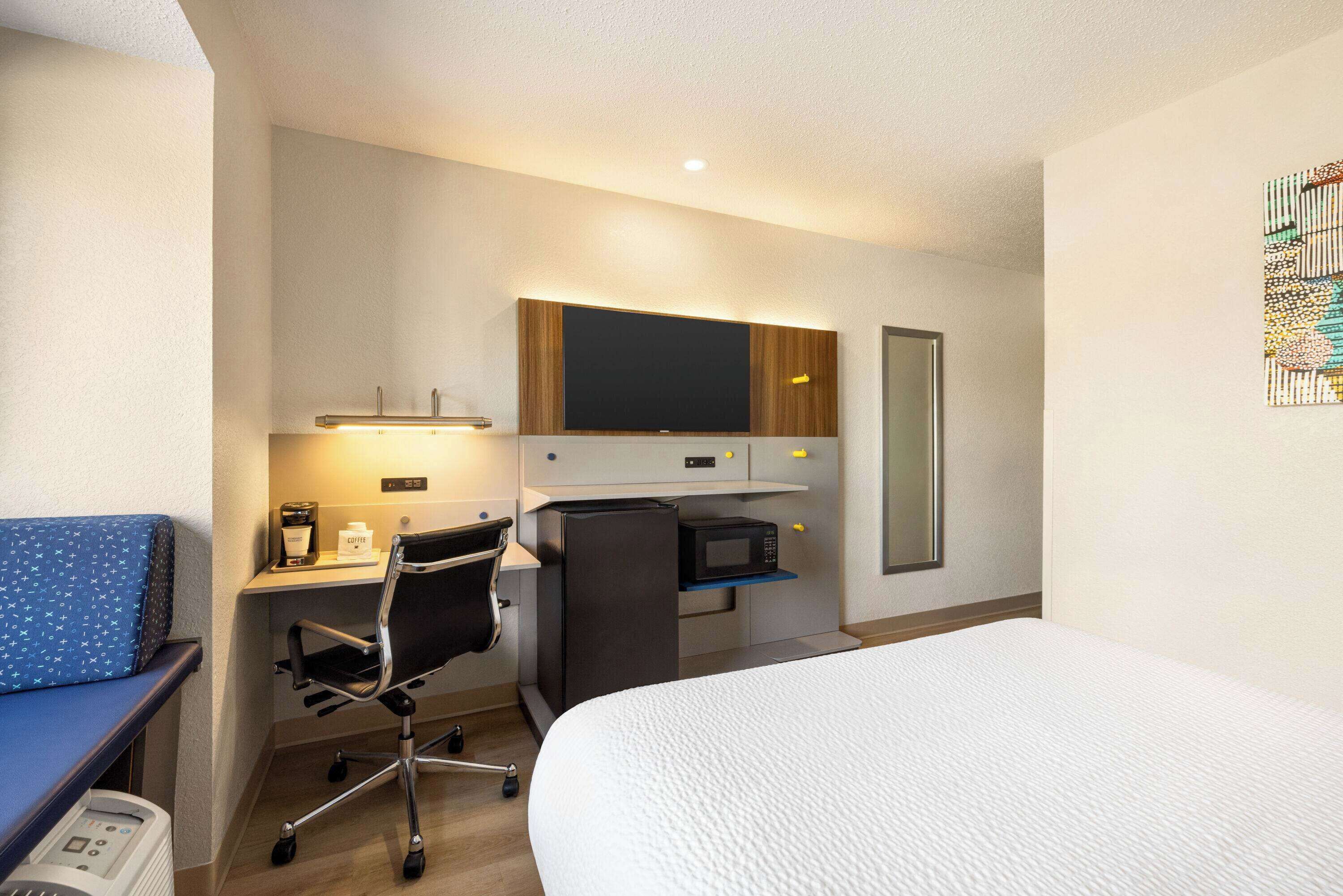 Habitació d'hotel amb un llit, una taula de treball amb un ordinador, una cadira, un microones i una televisió.