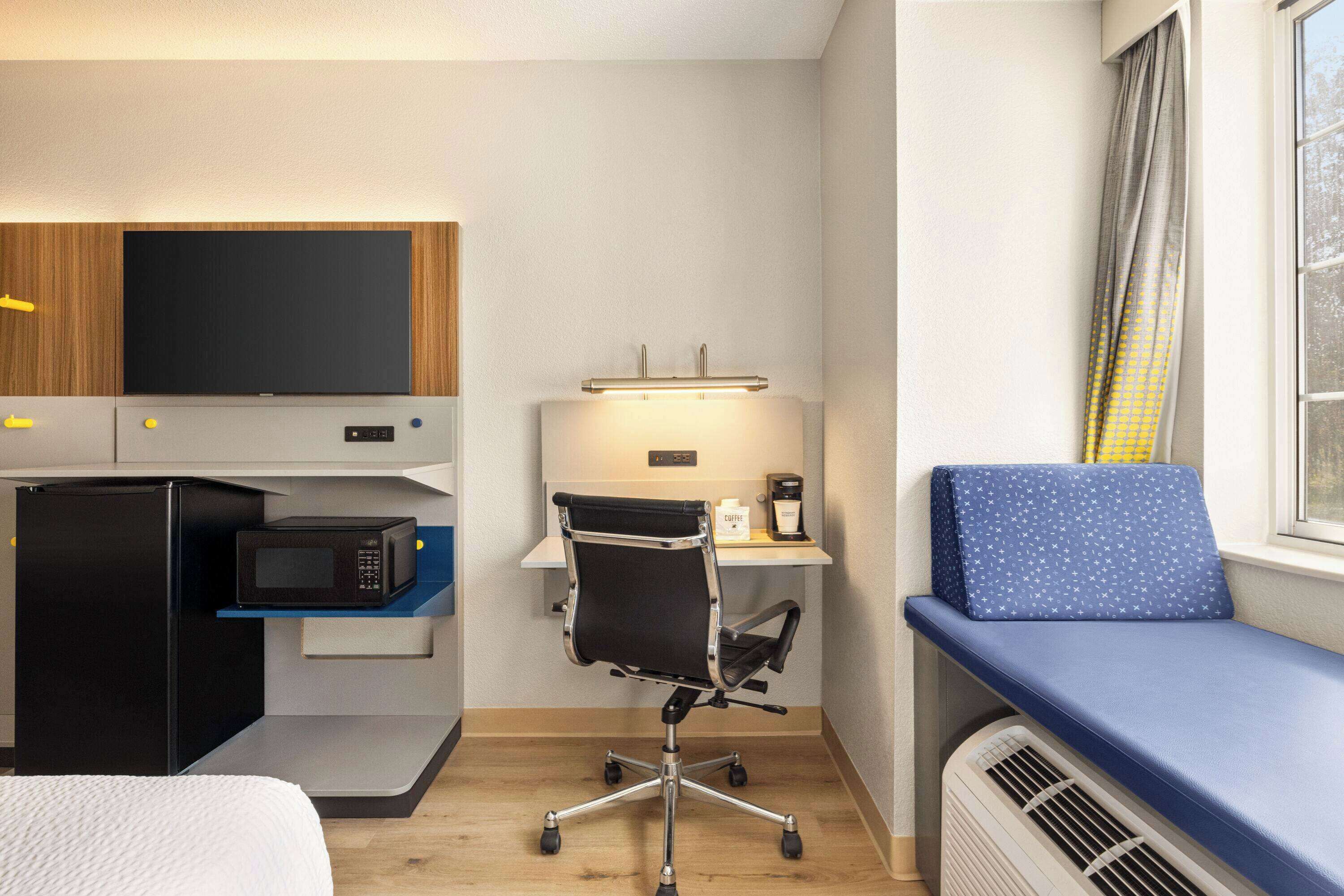 A compact hotel room with a kitchenette, a desk with a chair, a bed, and a window with curtains.