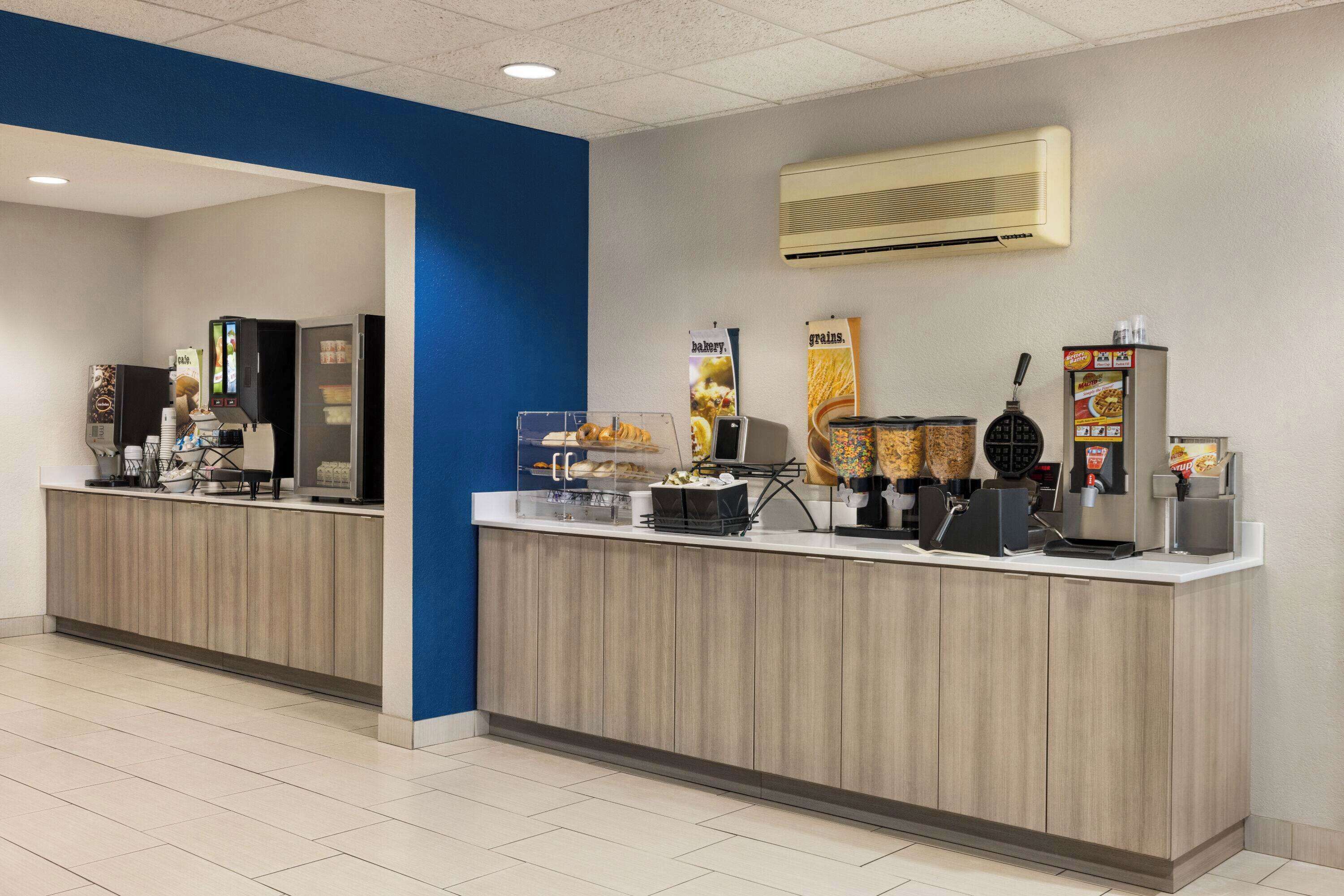 Un área de desayuno autoservicio limpia y moderna, con una máquina de café, dispensador de cereales y diversas opciones de alimentos.