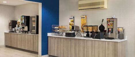 Un área de desayuno autoservicio limpia y moderna, con una máquina de café, dispensador de cereales y diversas opciones de alimentos.