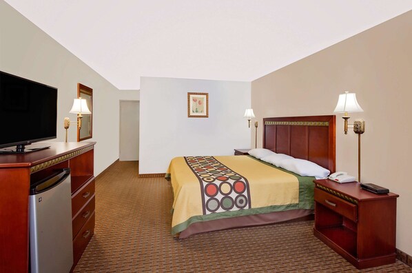 Una habitación de hotel con cama, televisión, escritorio con lámpara y un refrigerador pequeño.