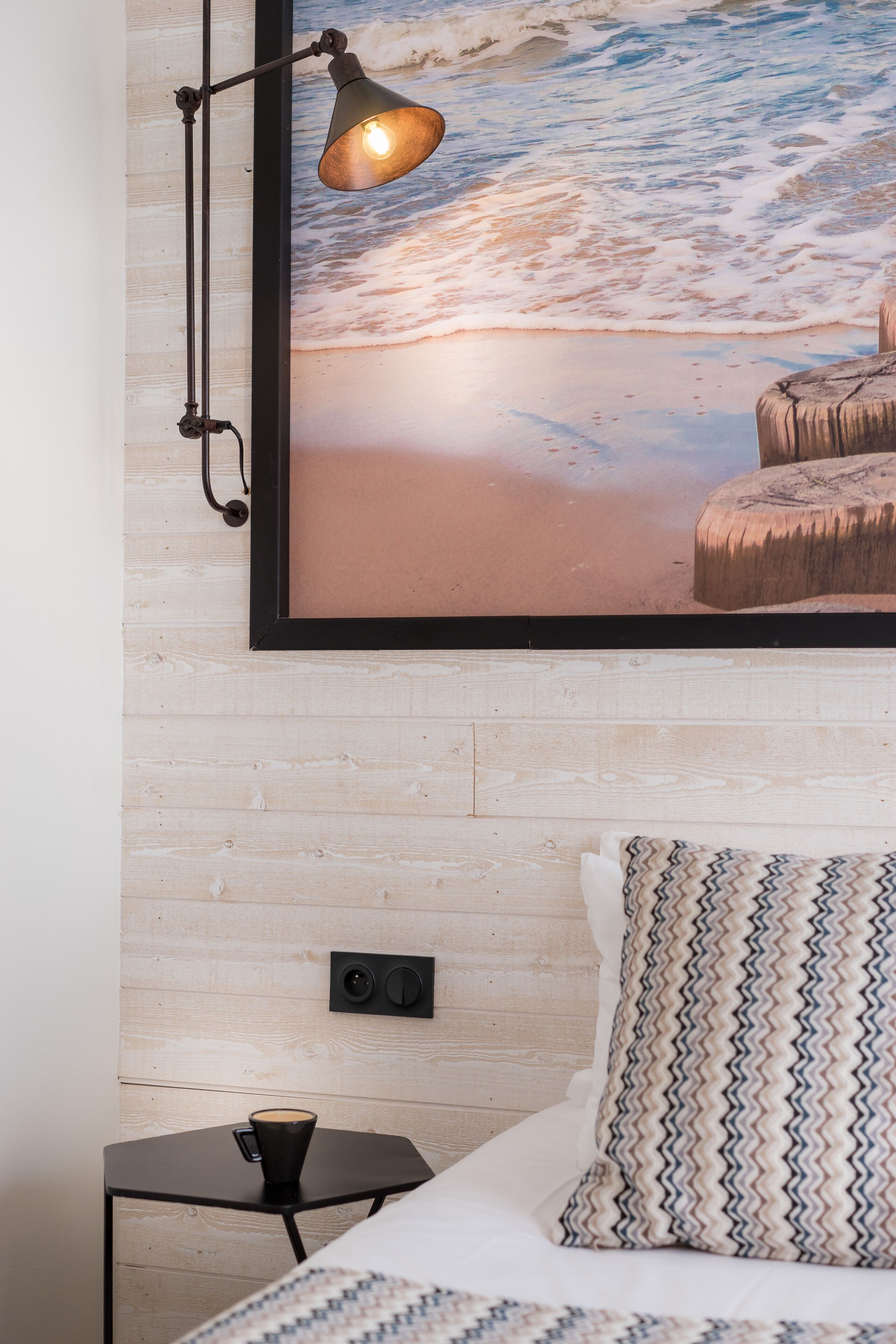 A bedroom with a wooden wall, a framed beach mural, a bedside lamp, a small table with a cup, and patterned pillows.