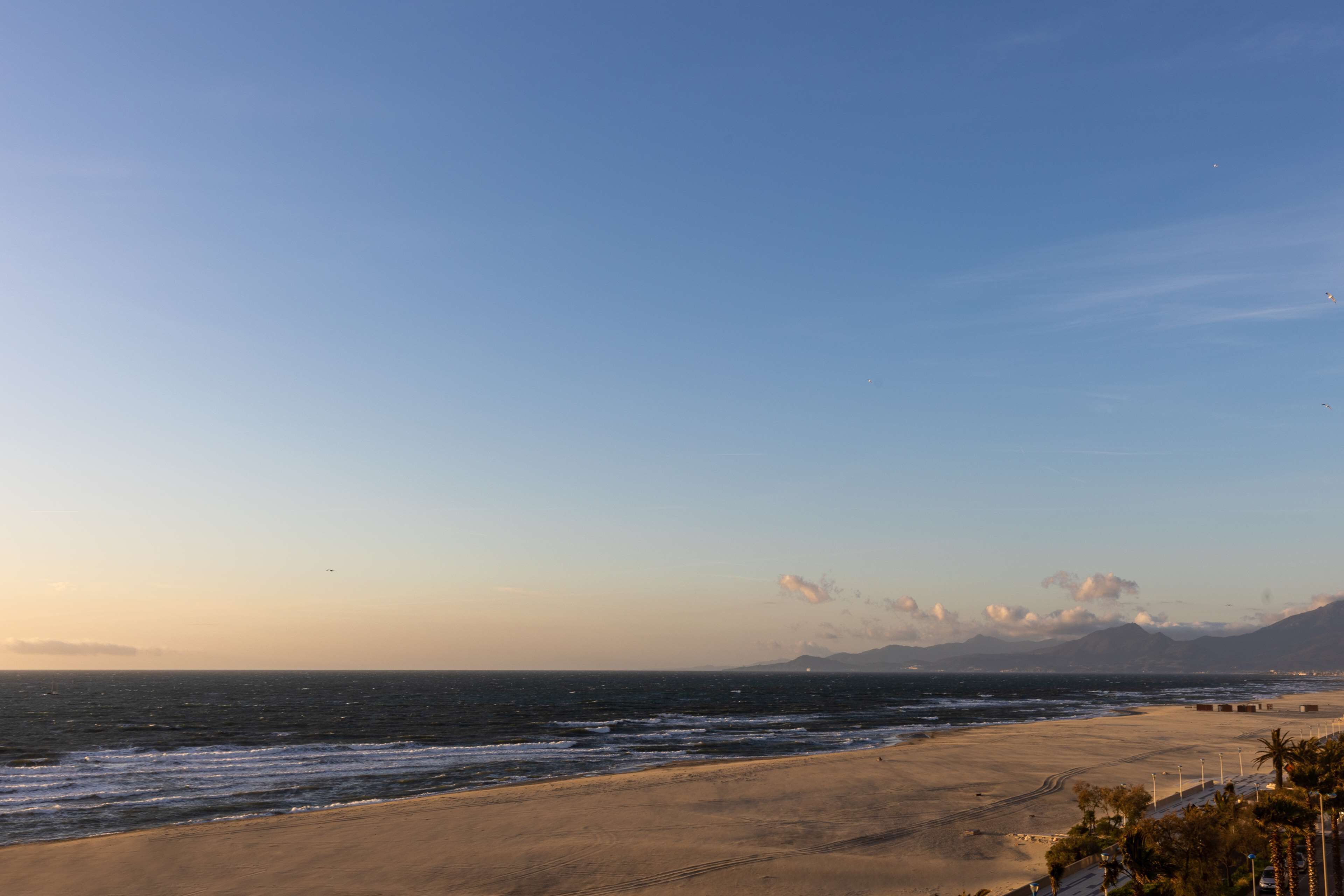 A beach with a clear sky, ocean waves, and distant mountains.