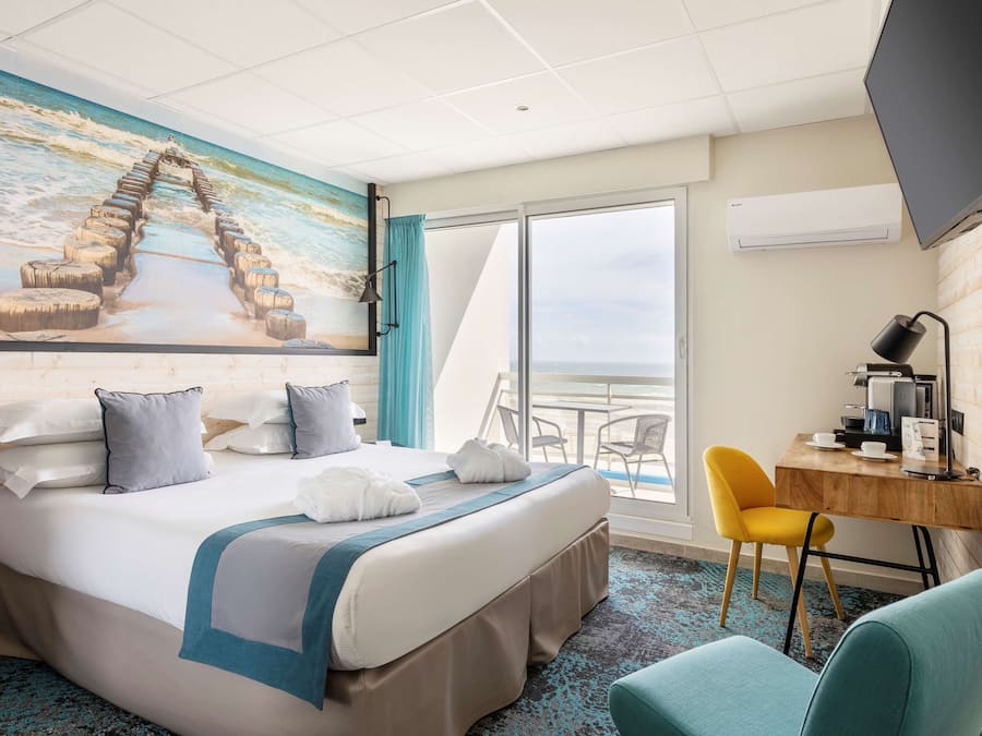 Quarto de hotel com cama, escrivaninha, cadeira, televisão e uma janela grande com vista para o oceano.
