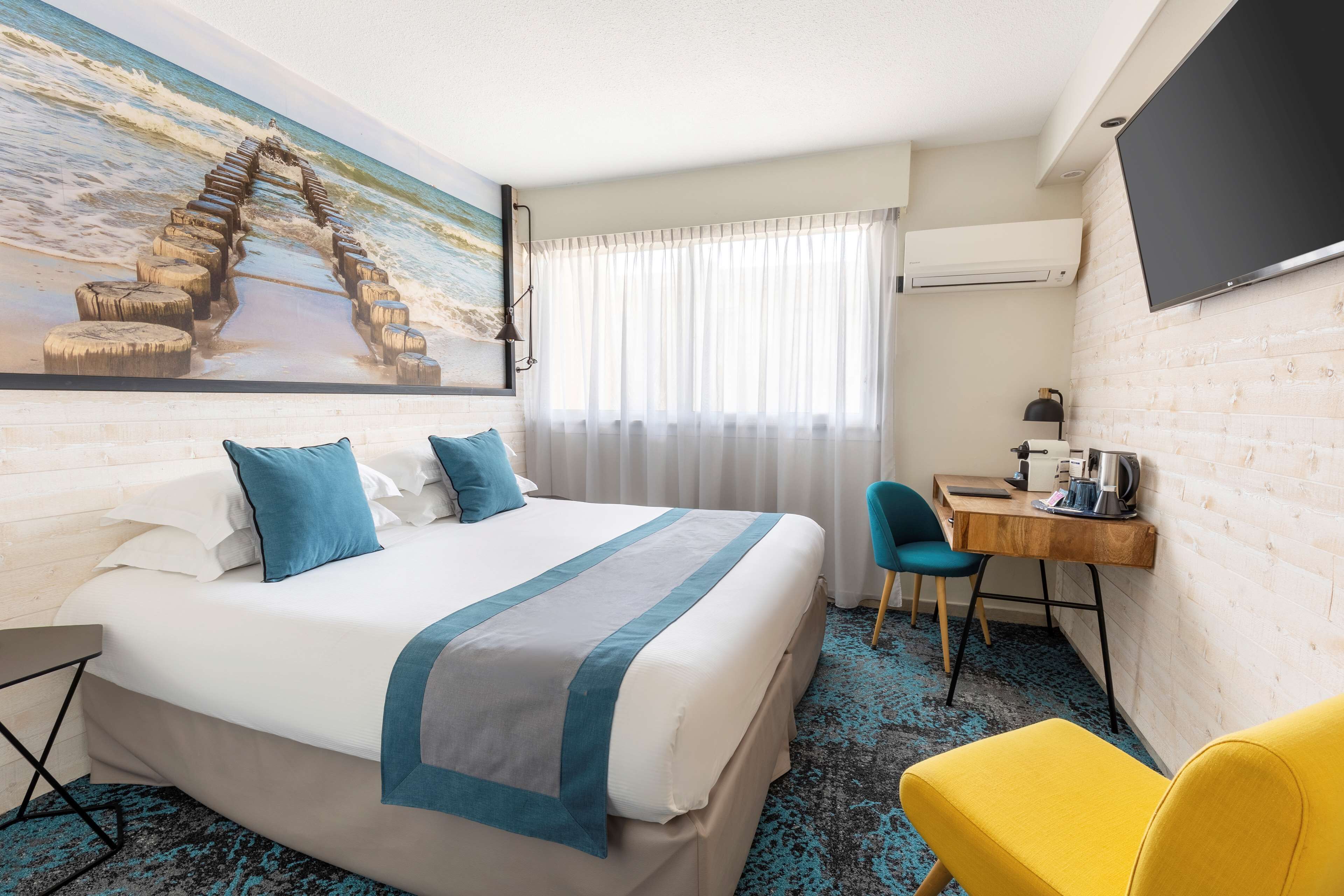 A hotel room with a bed, a desk, a chair, a TV, and a painting of a coastal scene.