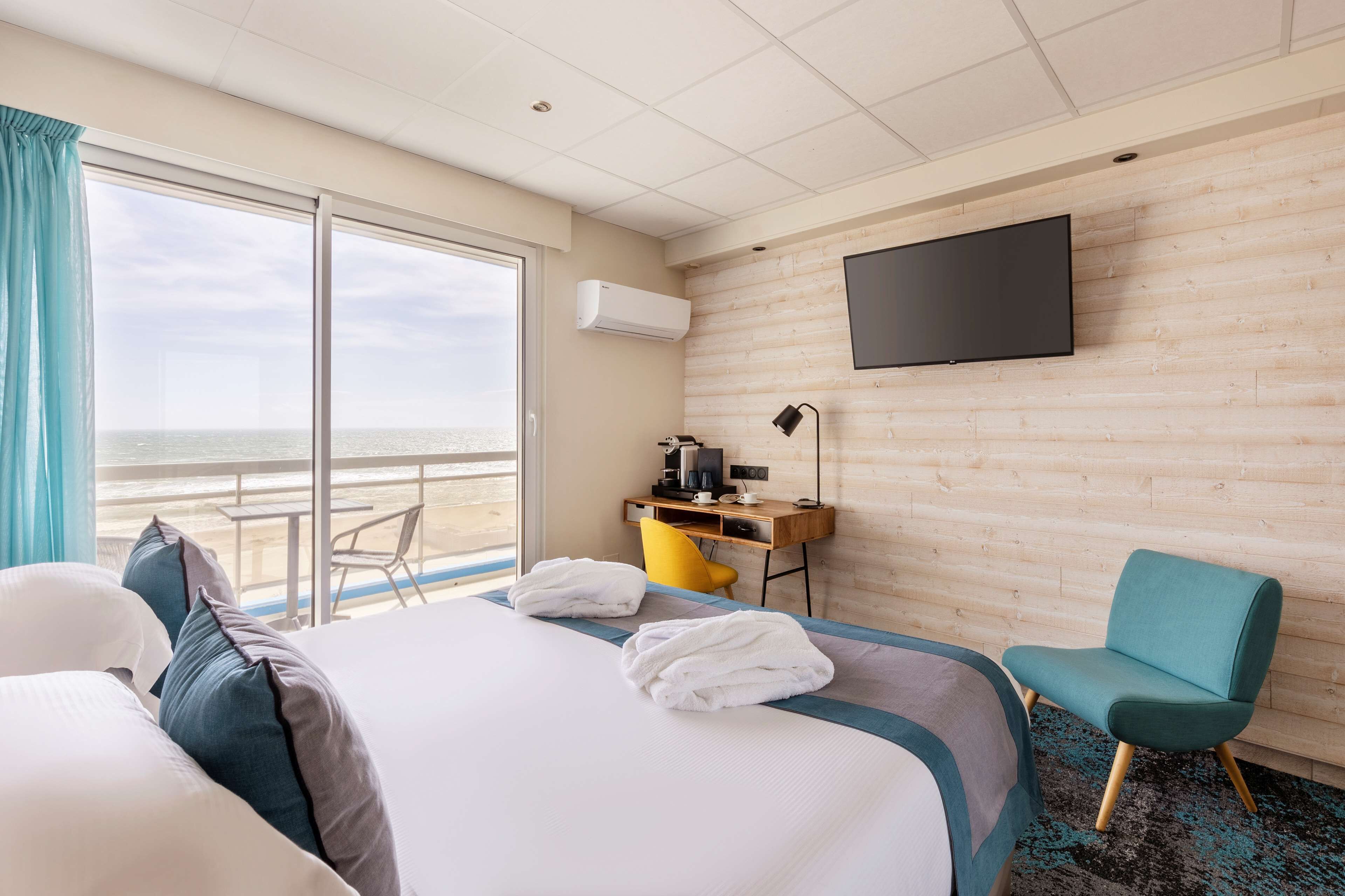 A hotel room with a large bed, a desk with a chair, a TV on the wall, and a view of the ocean.