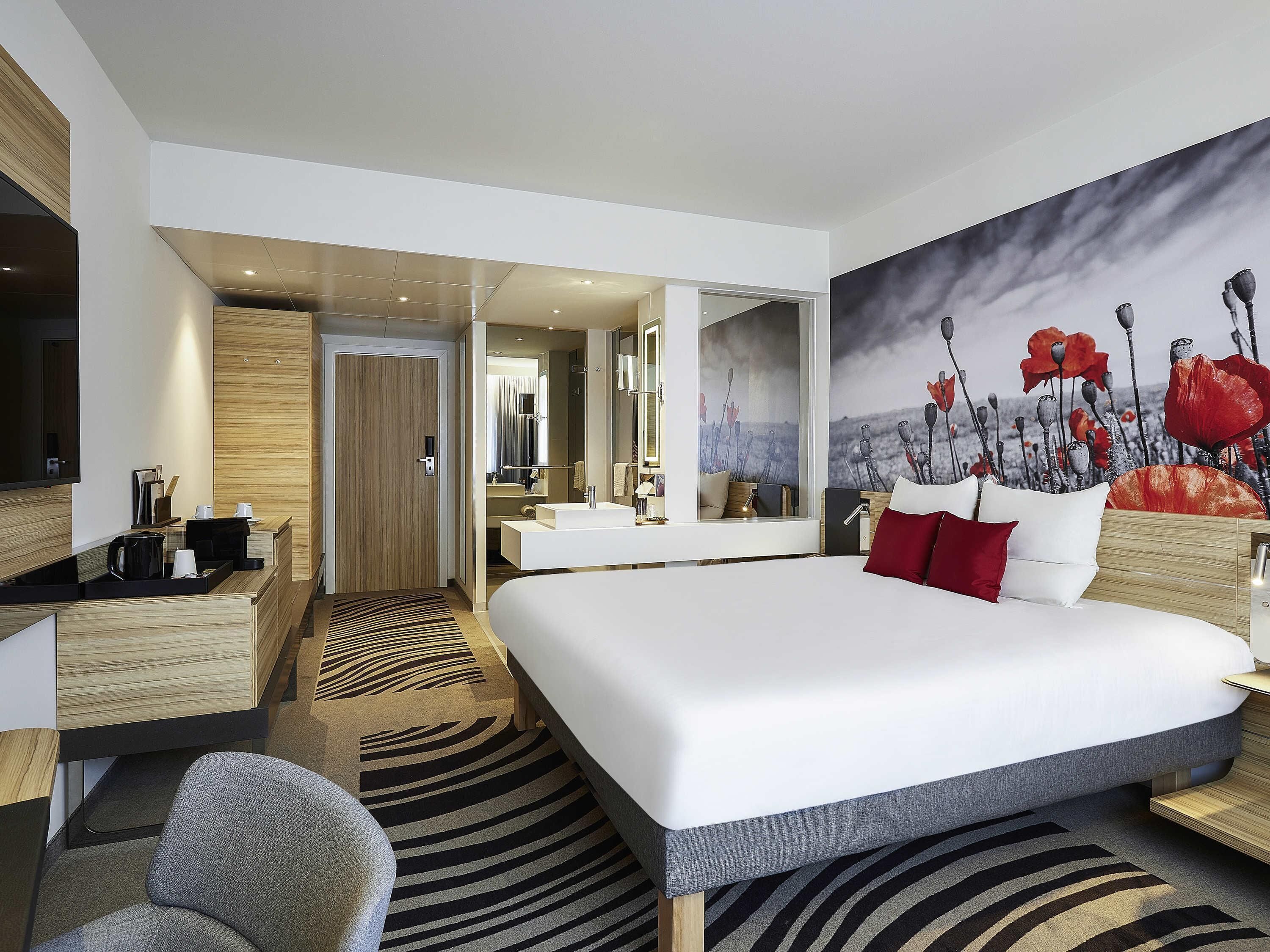 A modern hotel room with a large bed, a desk, a chair, a bathroom with a mirror, and a painting on the wall.