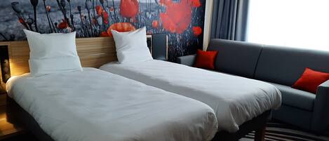Habitación de hotel con dos camas, un sofá gris y un ventanal con cortinas.