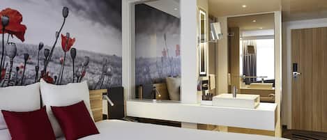 A modern hotel room with a large bed, a bedside table, a mirror, and a view of the bathroom.