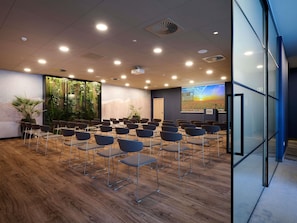 Un salón de conferencias con filas de sillas, una pantalla de proyector y un amplio ventanal con vistas a zonas verdes.