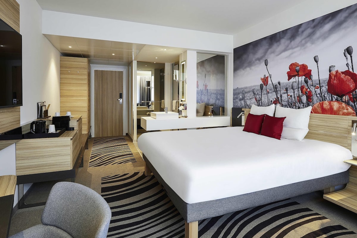 A modern hotel room with a large bed, a desk, a chair, a bathroom with a mirror, and a painting on the wall.