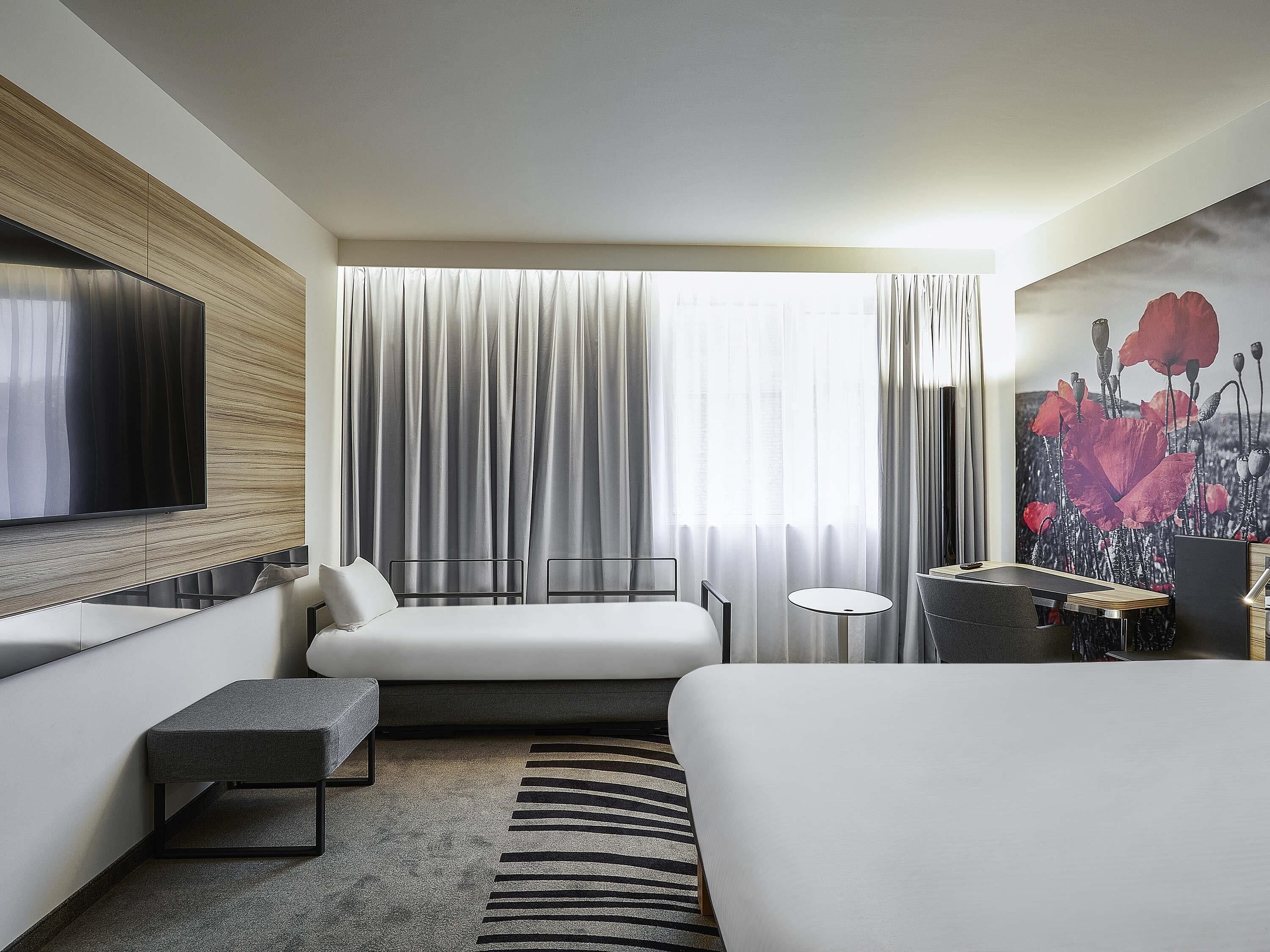 A modern hotel room with a large bed, a sofa, a small table, a TV mounted on the wall, and a window with curtains.