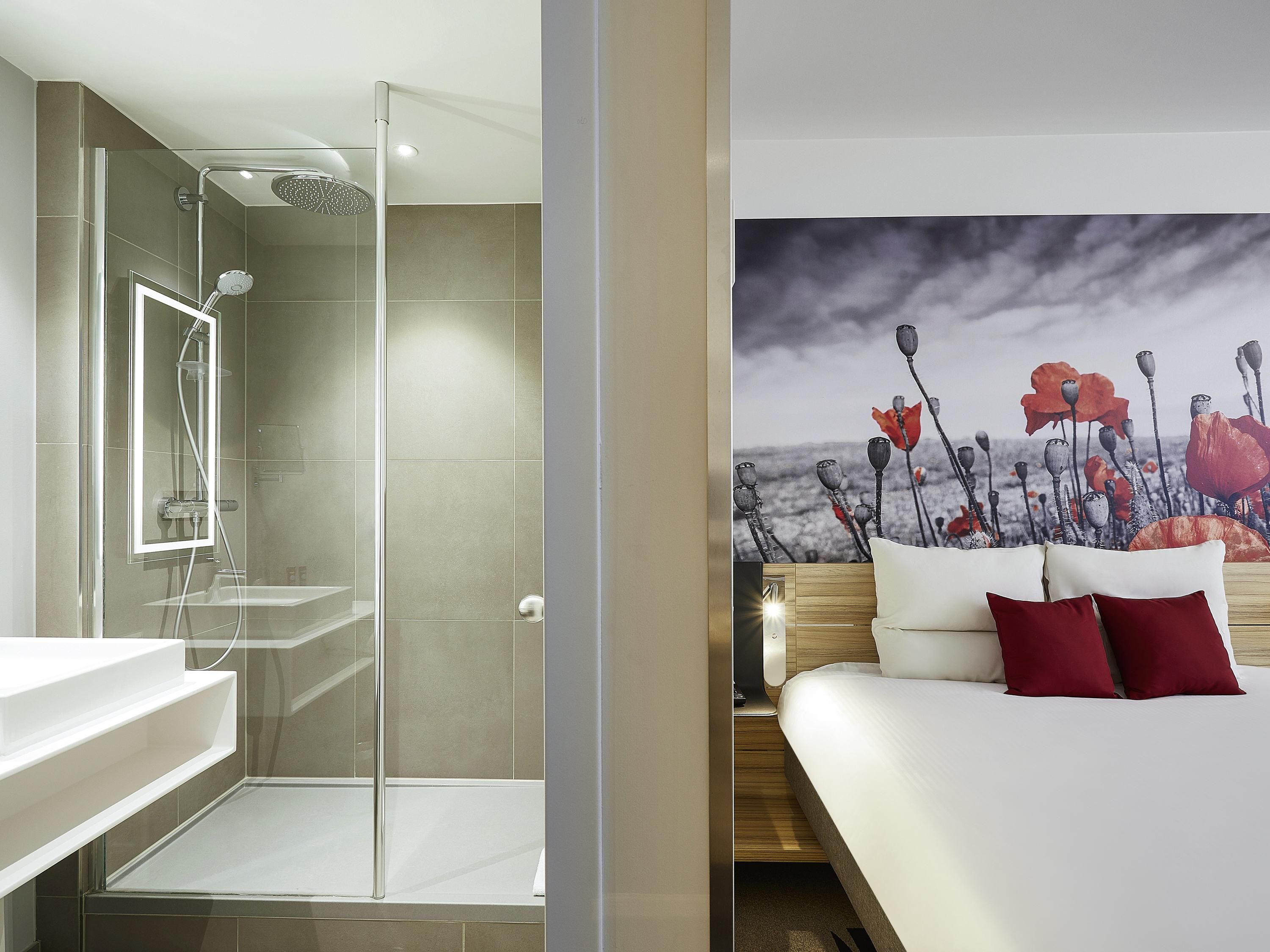 A modern bathroom with a glass shower enclosure, a white sink, and a wooden bedside table with a white mattress and red pillows.