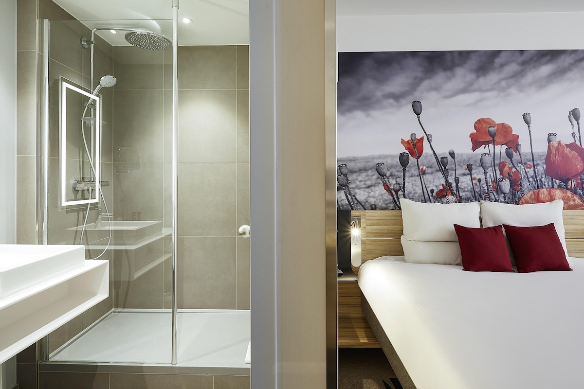 Un baño moderno con cabina de ducha de vidrio, un lavamanos blanco y una mesita de noche de madera con colchón blanco y almohadas rojas.