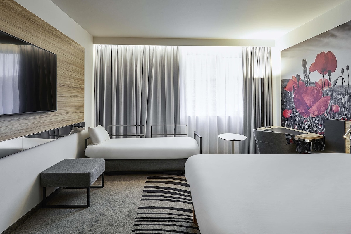 Una habitación de hotel moderna con una cama grande, un sofá, una mesita y un televisor montado en la pared, además de una ventana con cortinas.