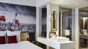 A modern hotel room with a large bed, a white vanity with a mirror, and a view of the sea through the window.