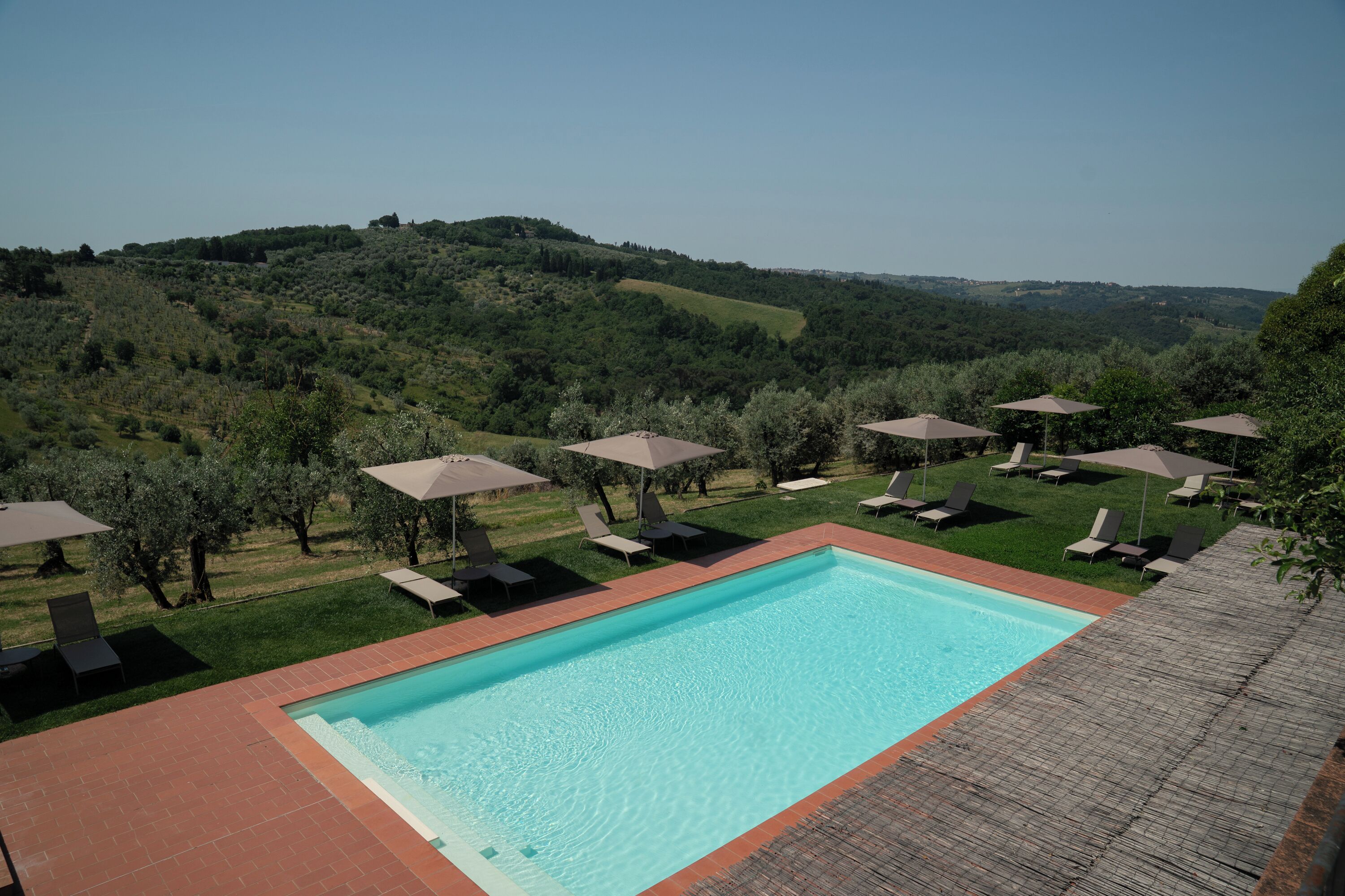 A swimming pool with lounge chairs and umbrellas, surrounded by a grassy area and trees, with a view of rolling hills in the background.