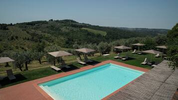 A swimming pool with lounge chairs and umbrellas, surrounded by a grassy area and trees, with a view of rolling hills in the background.
