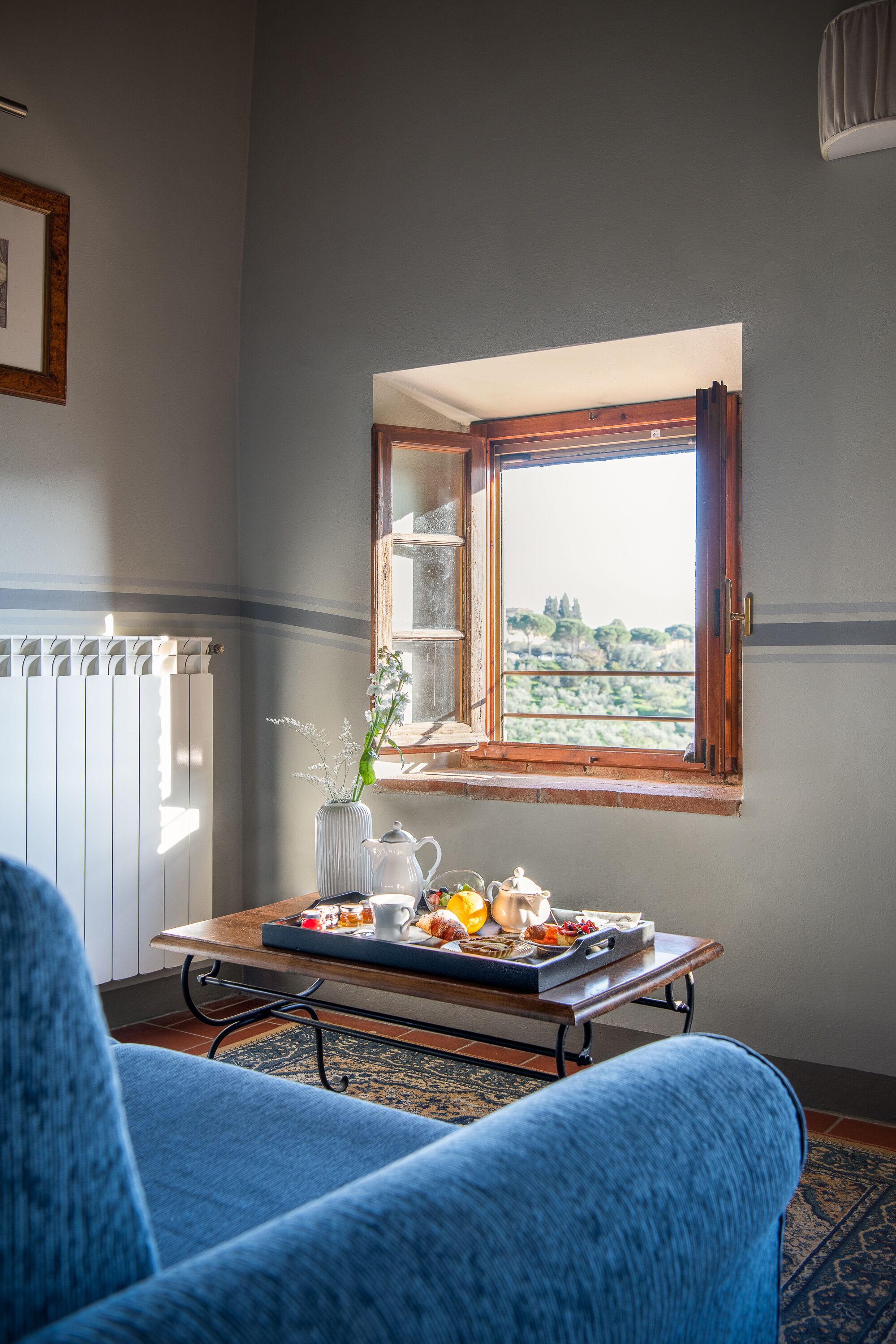 Una habitación con vistas a través de una ventana, una mesa con una bandeja de comida, un radiador y una silla azul.