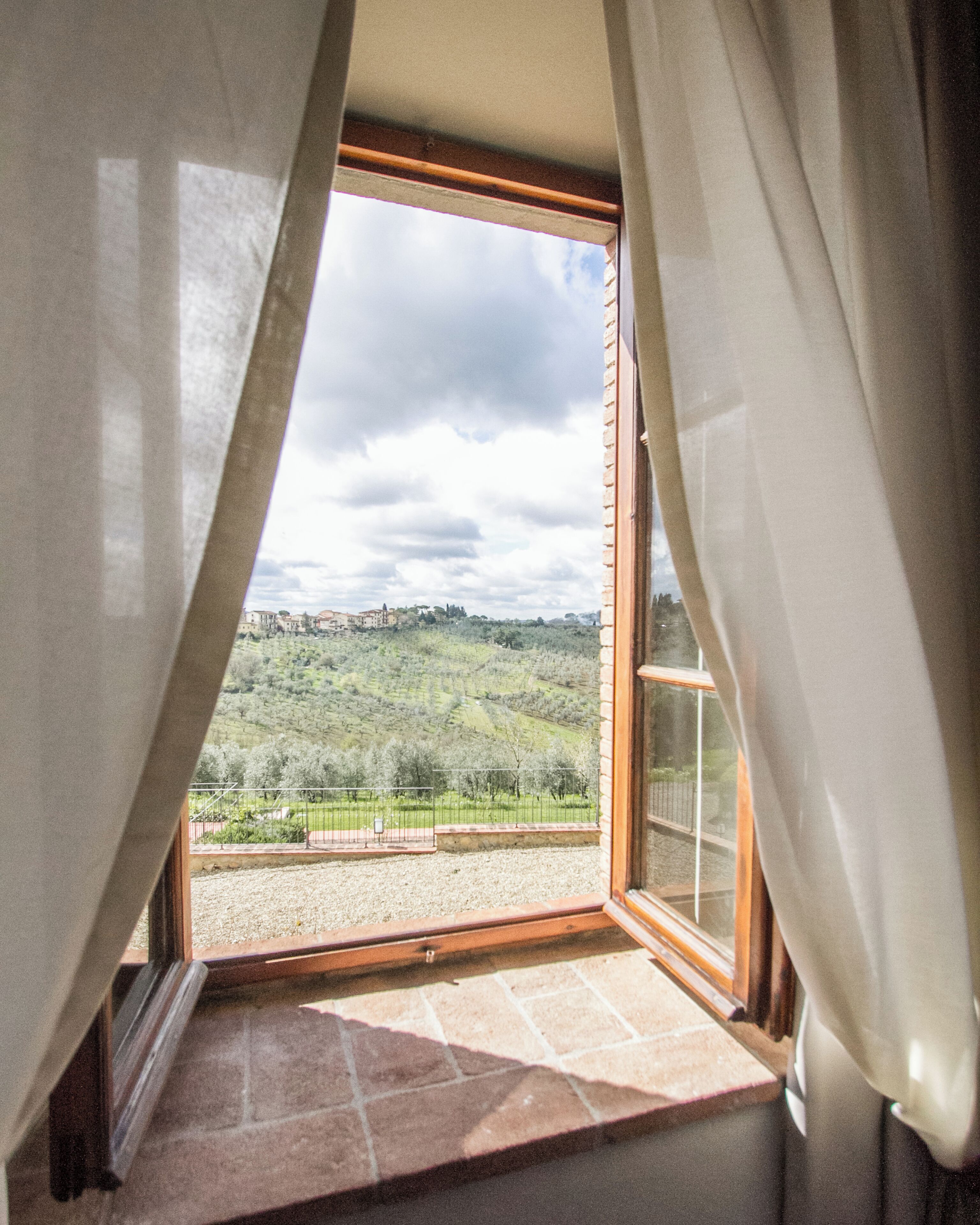 A room with a large window and curtains, offering a view of a green landscape and distant buildings.
