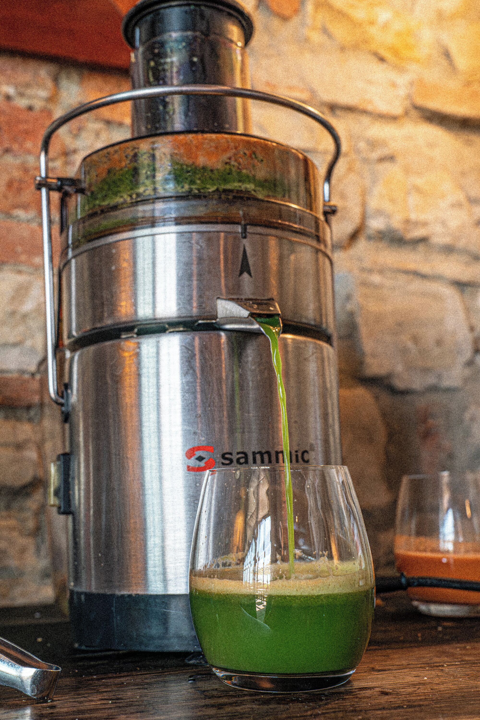 Un dispensador metálico de agua con un vaso de líquido verde, una pared de piedra de fondo y un recipiente metálico a su lado.