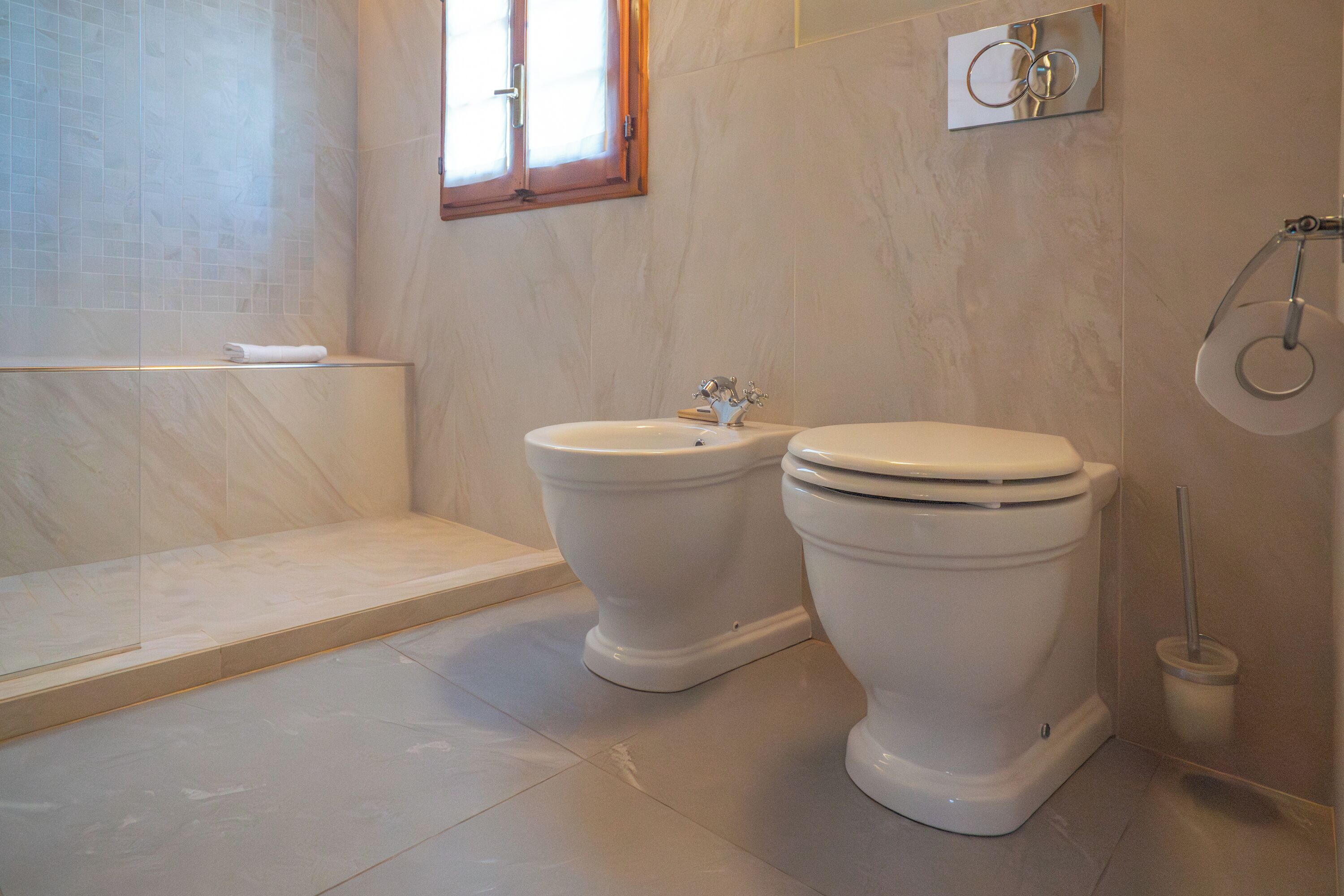 Ein Badezimmer mit zwei Toiletten, einem Bidet, einem Fenster und einem Toilettenpapierhalter.