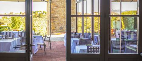 An outdoor dining area with tables set for a meal, large windows, and a stone wall in the background.