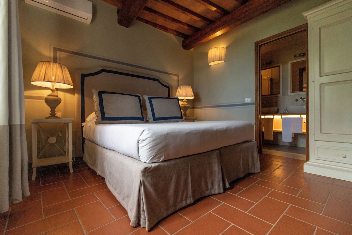 A bedroom with a large bed, two bedside tables with lamps, a wooden ceiling, and a bathroom visible through an open door.