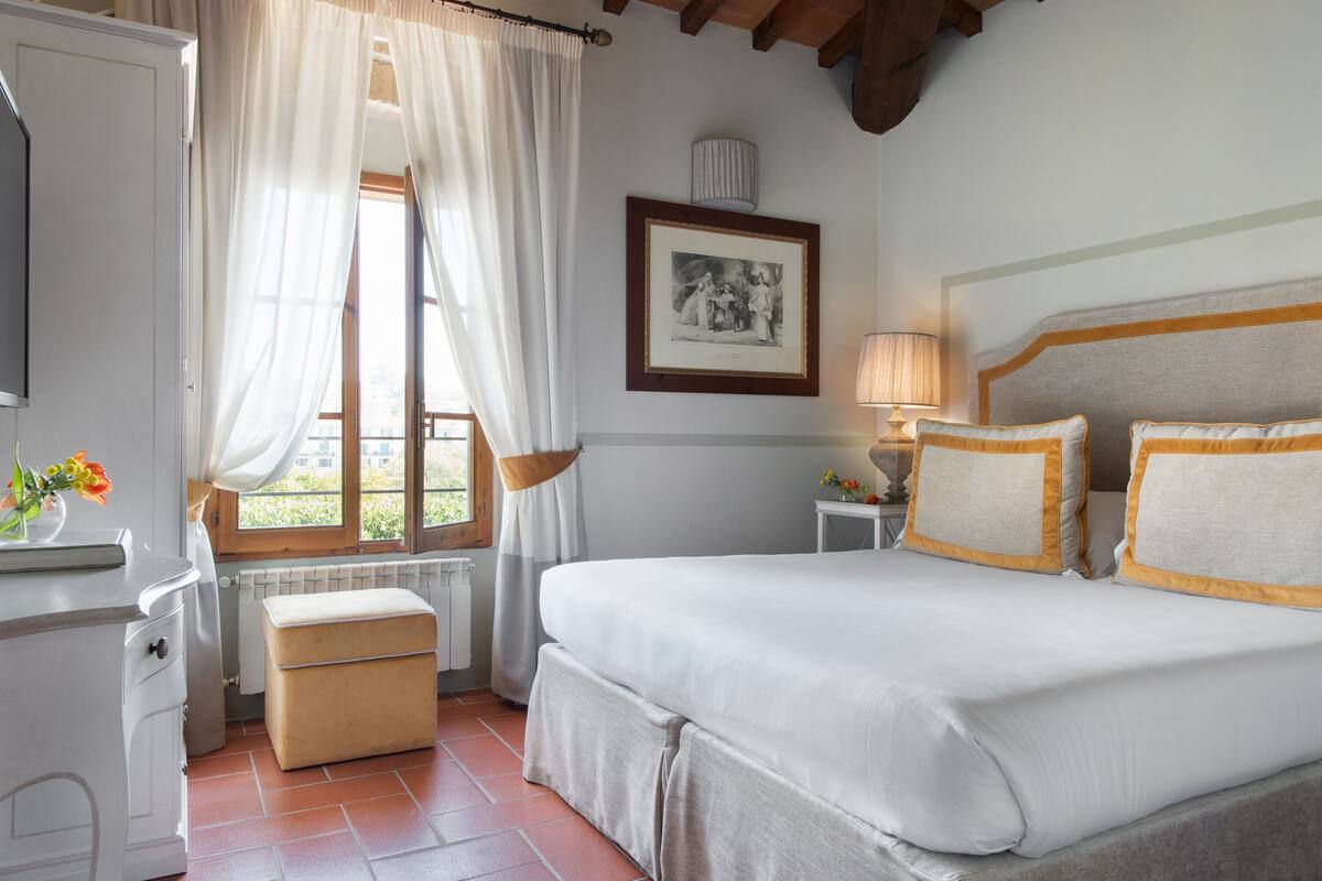 A bedroom with a bed, bedside table, lamp, window with curtains, radiator, and a framed picture on the wall.