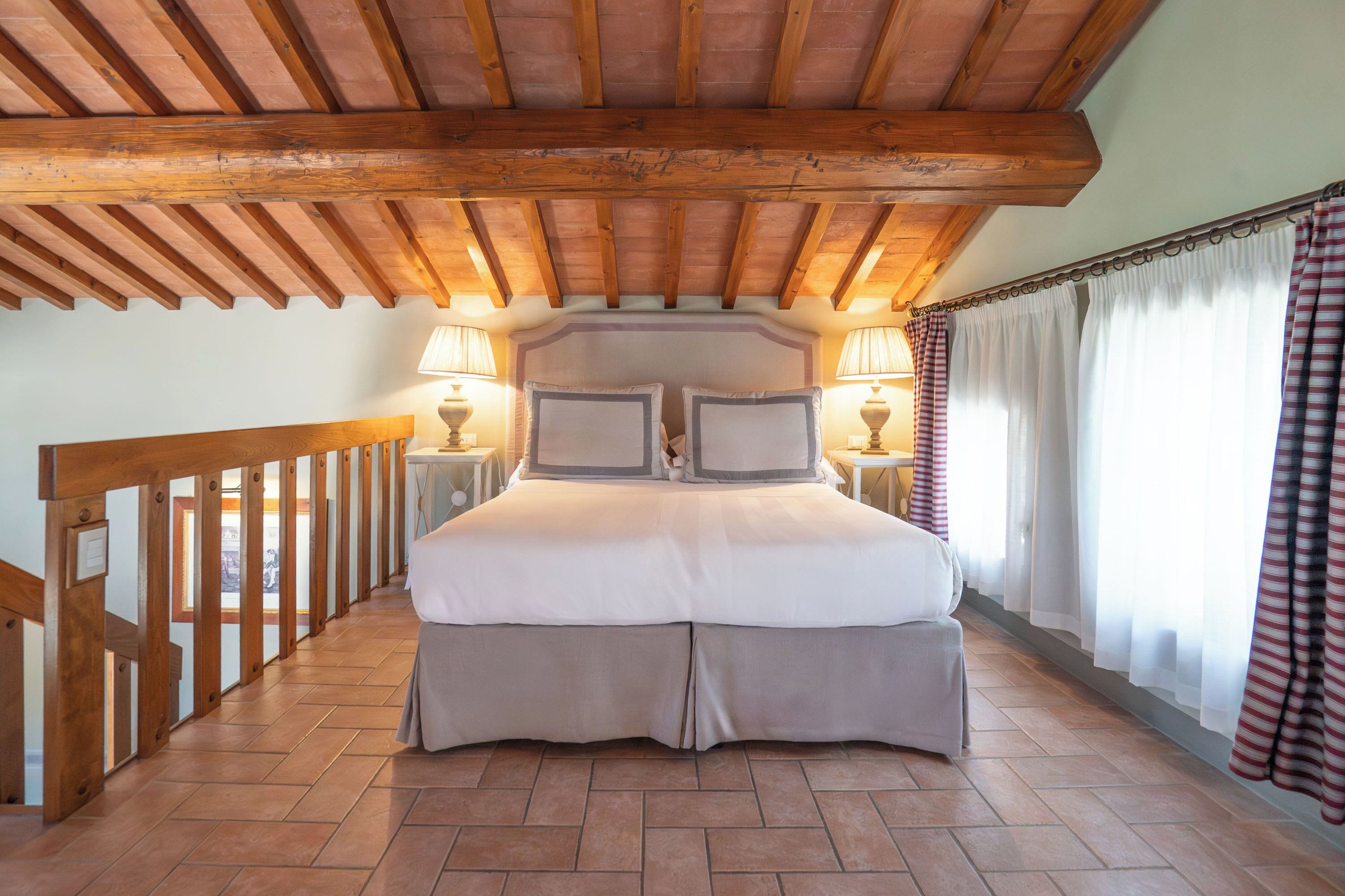 Une chambre avec un plafond en bois, un lit recouvert d’une couette grise et blanche, deux lampes de chevet et une vue sur un balcon.