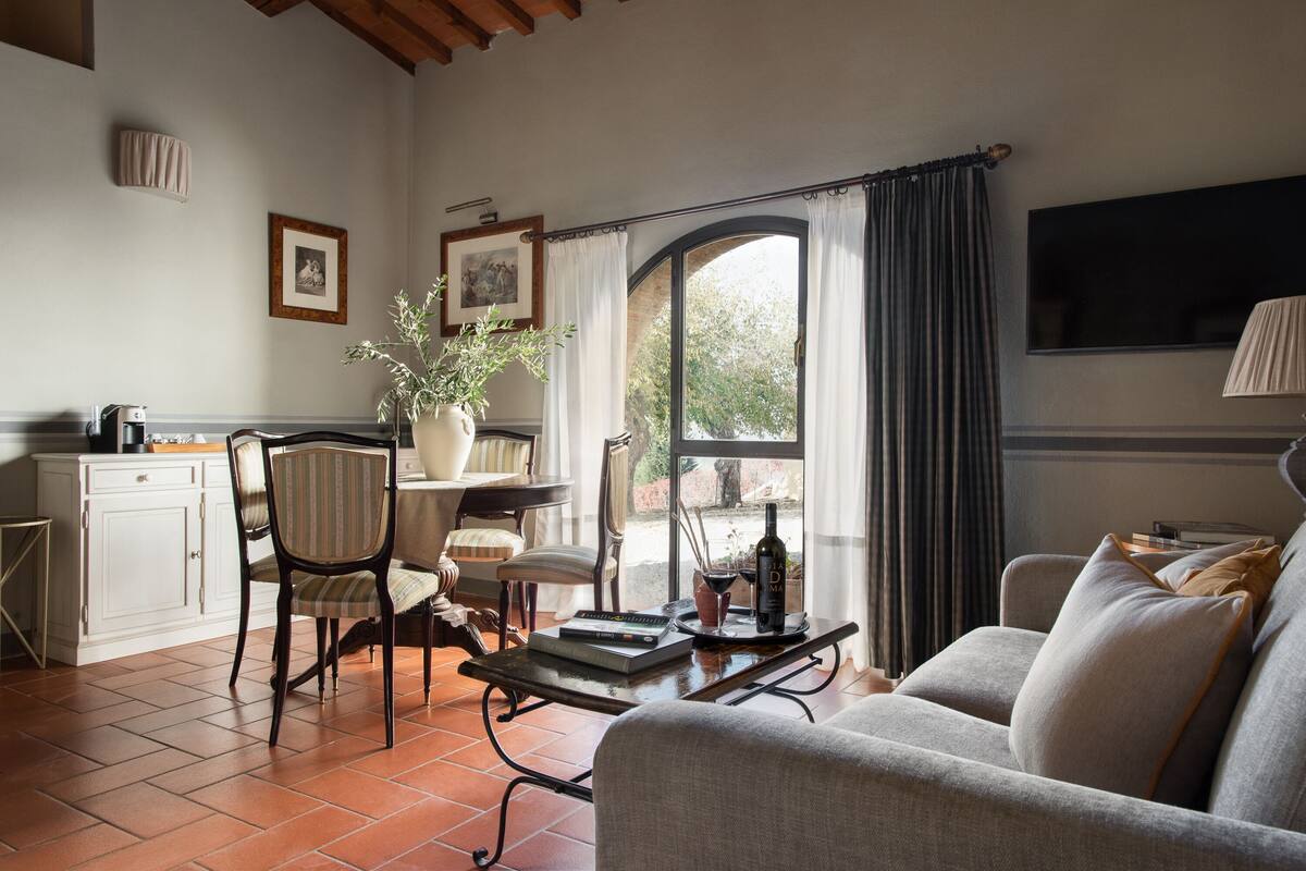 A living room with a sofa, chairs, a coffee table, a dining table, and a potted plant. There is a window with curtains, a television, and a fireplace.