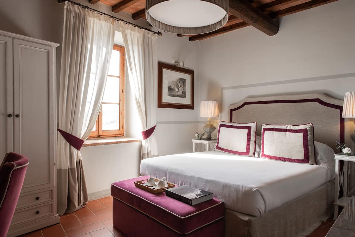 A bedroom with a large bed, a wooden ceiling, a window with curtains, a bedside table with a lamp, and a chair.