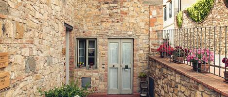 A stone building with a tiled courtyard, a large planter, and a metal railing with flowers.