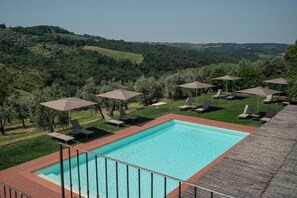 A rectangular swimming pool with a tiled border, surrounded by lounge chairs and umbrellas, set against a backdrop of rolling hills and trees.