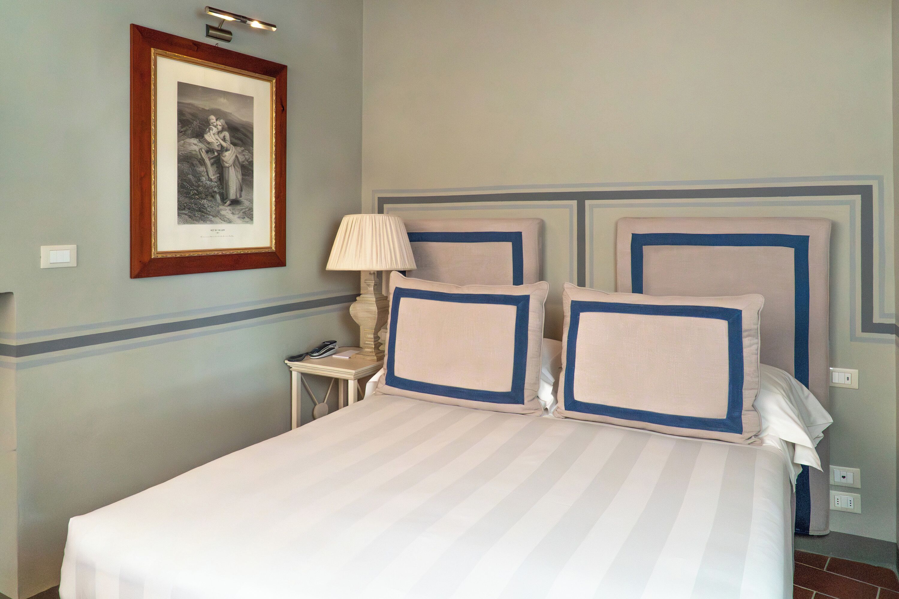 A hotel room with a bed, a nightstand, a lamp, and a framed picture on the wall.