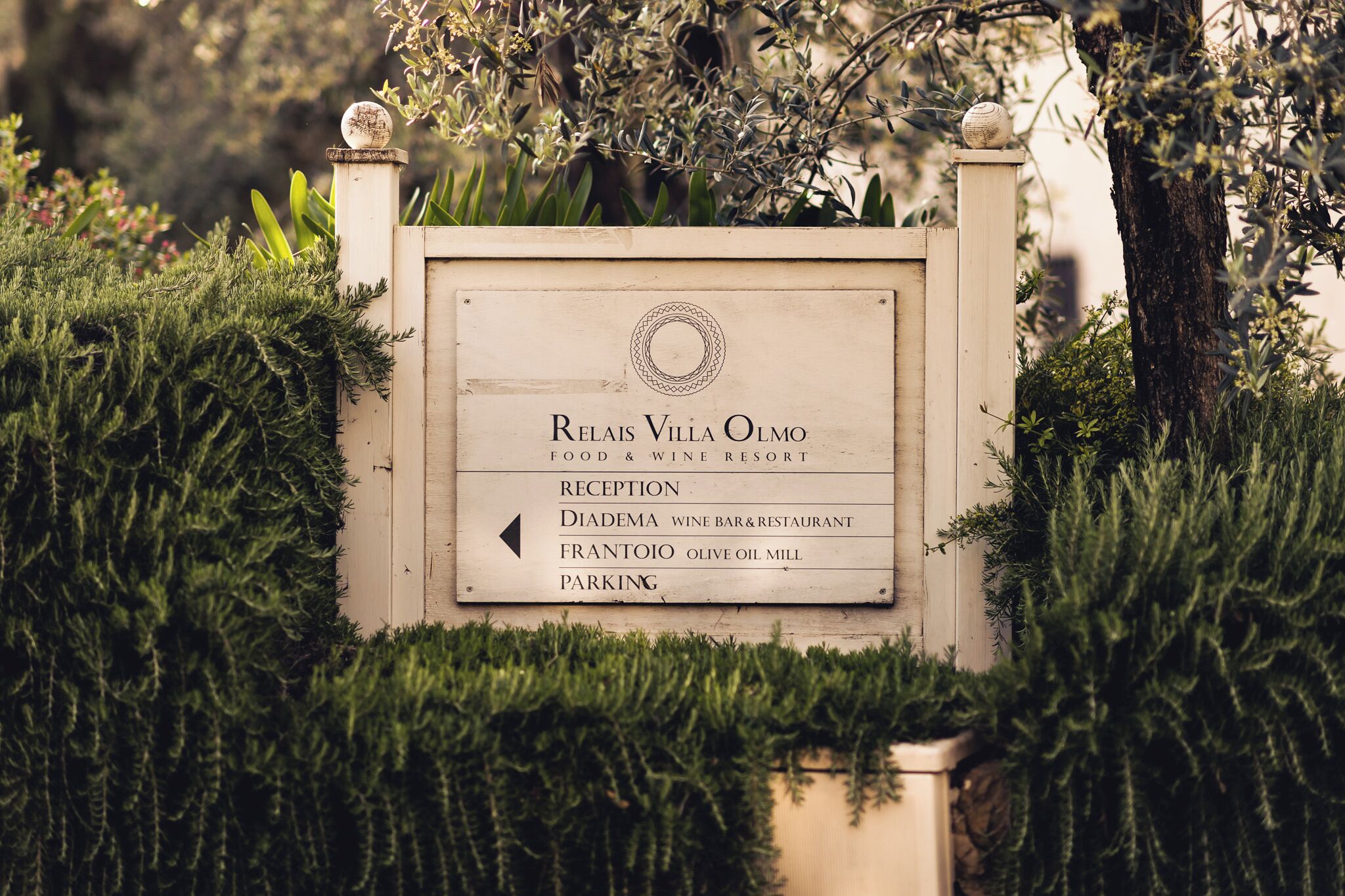A sign for Relais Villa Olmo, a food and wine resort, with directions to Reception, Diadema Wine Bar & Restaurant, Frantoio Olive Oil Mill, and Parking.