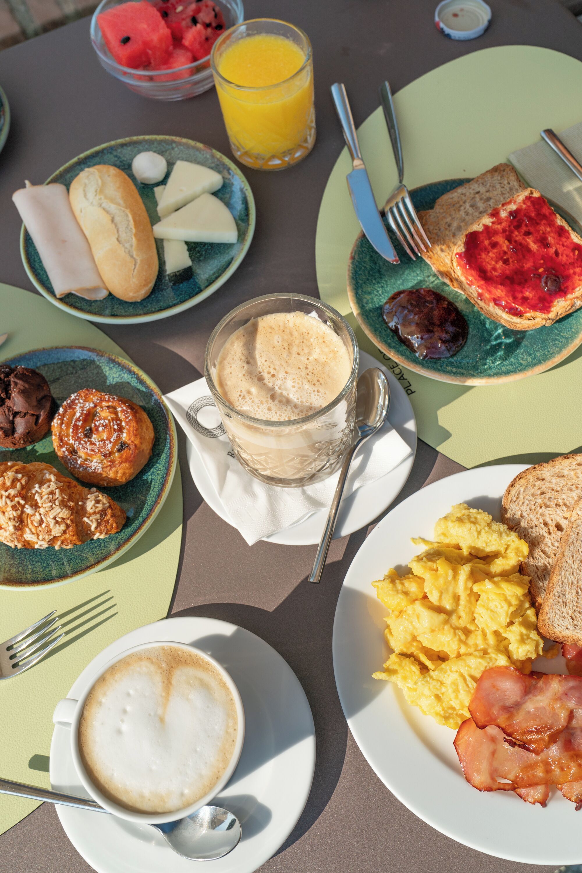 A breakfast table with various food items including toast, scrambled eggs, bacon, pastries, and a glass of orange juice.