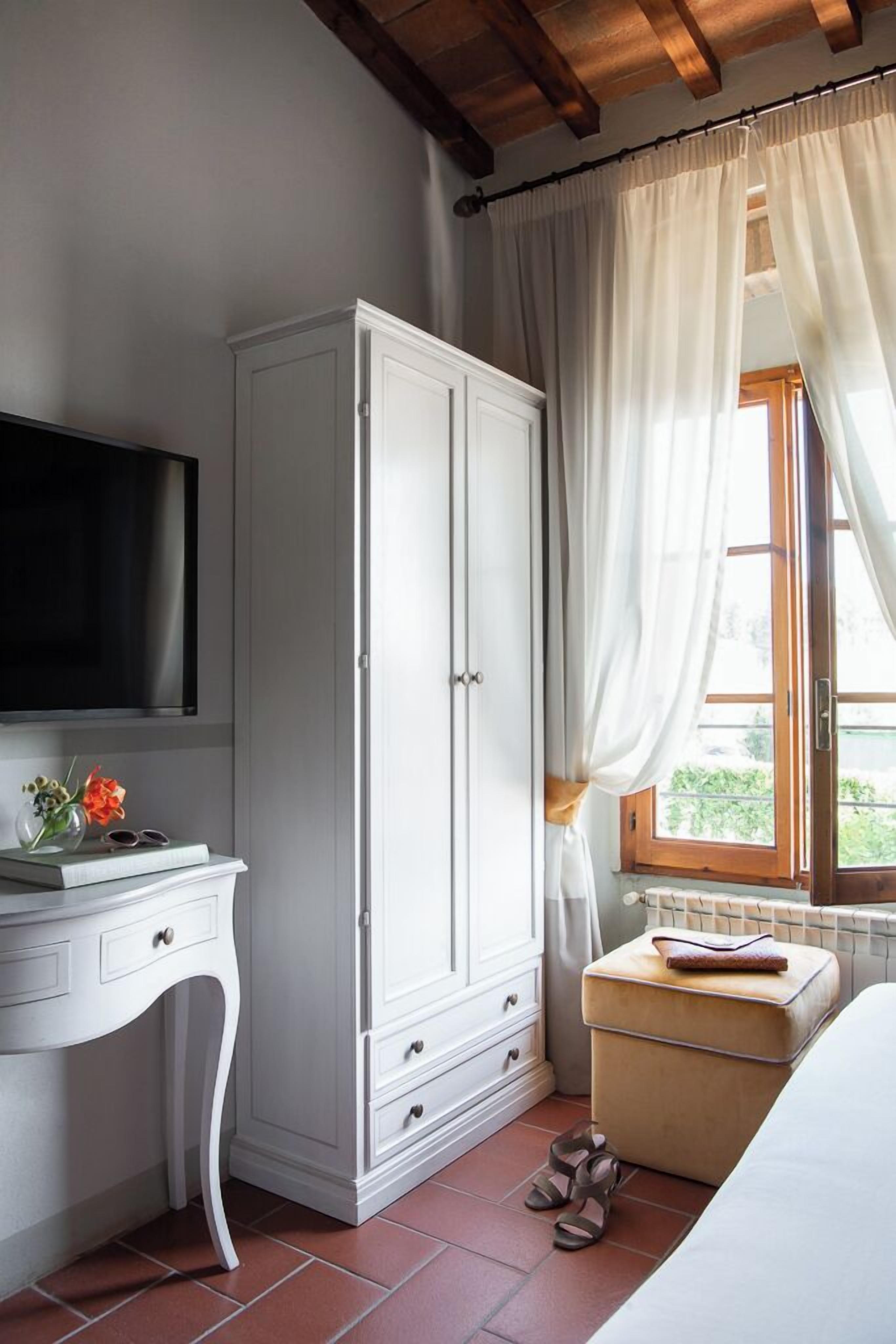 A bedroom with a white wardrobe, a bedside table with a TV, a window with curtains, and a small bedside table with a book and slippers.