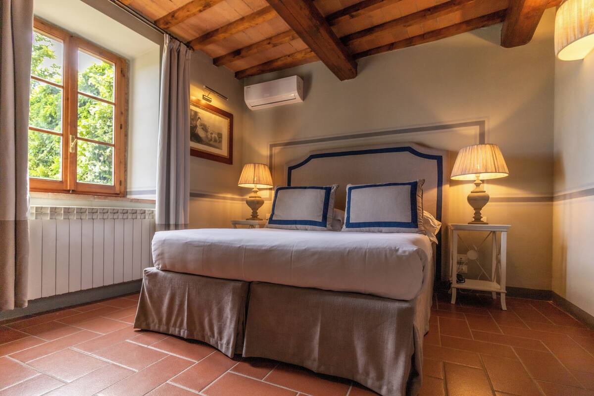A bedroom with a large bed, two bedside lamps, a window with curtains, and a radiator.