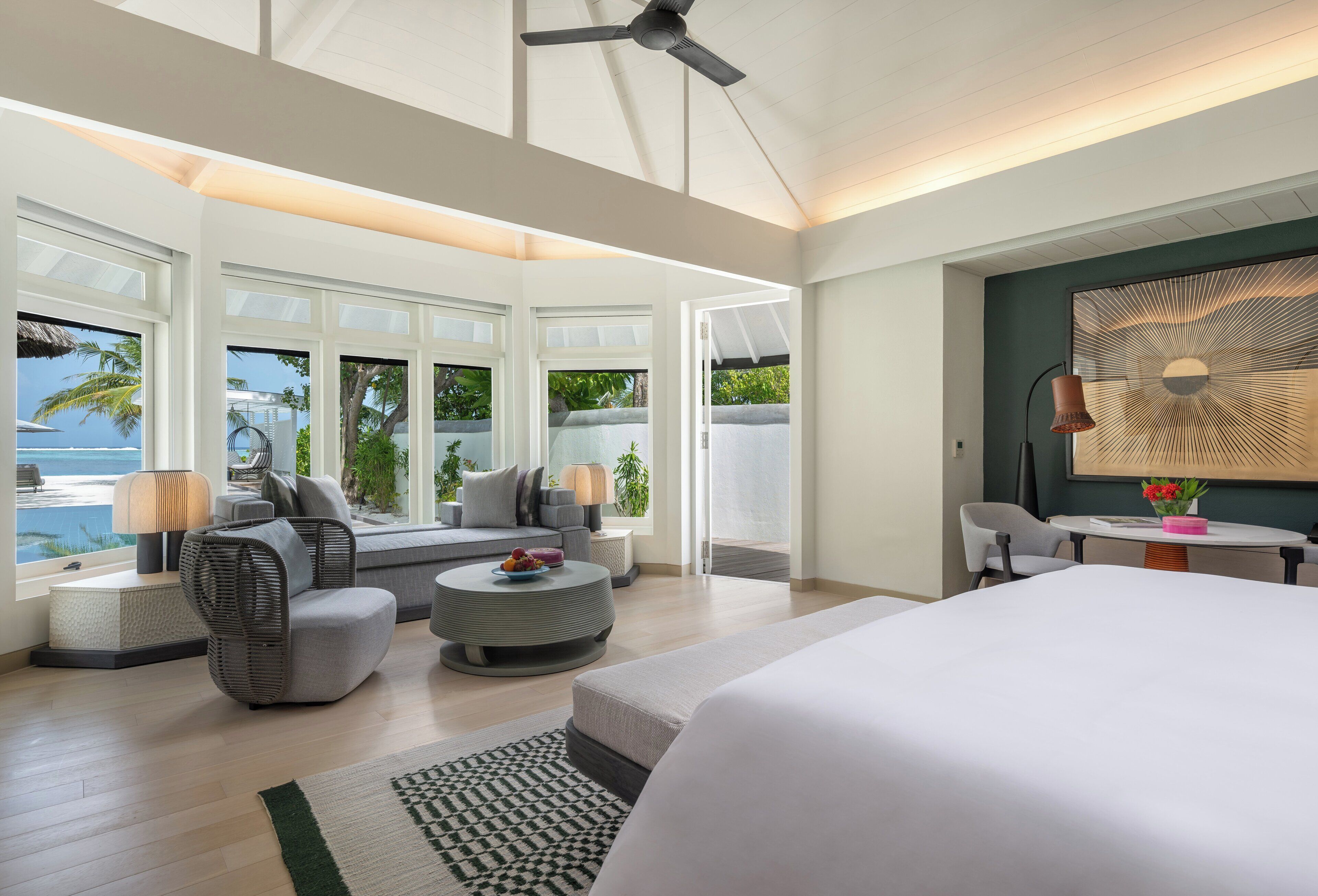 A modern hotel room with a large bed, a seating area with a sofa and armchairs, a dining table, and large windows offering a view of the ocean.