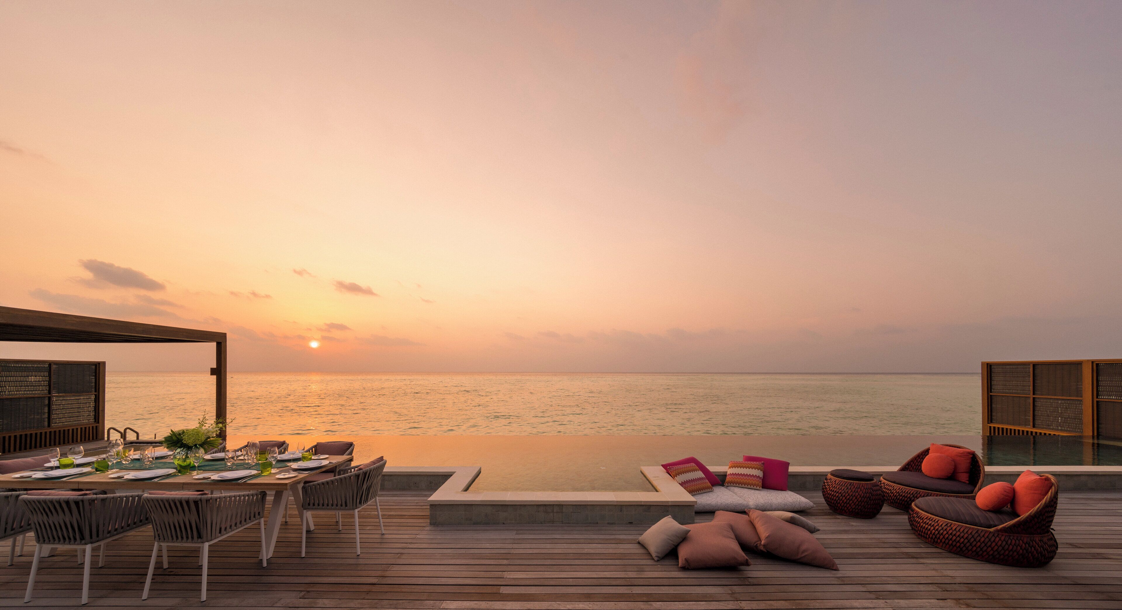 An outdoor dining area with a view of the sea at sunset, wooden deck, and wicker furniture.