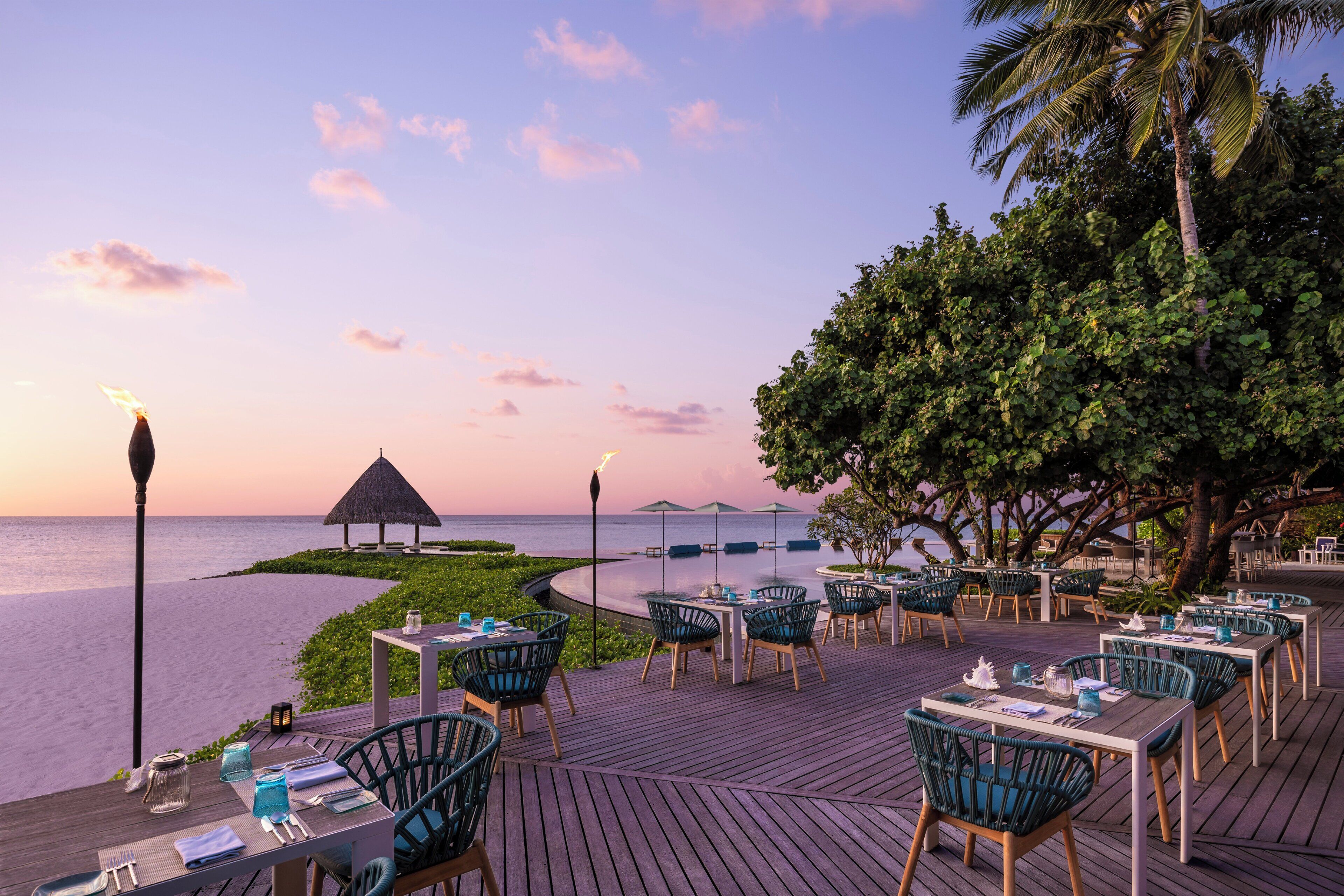 Un área de comedor al aire libre con mesas y sillas, con vista a una playa y al océano.