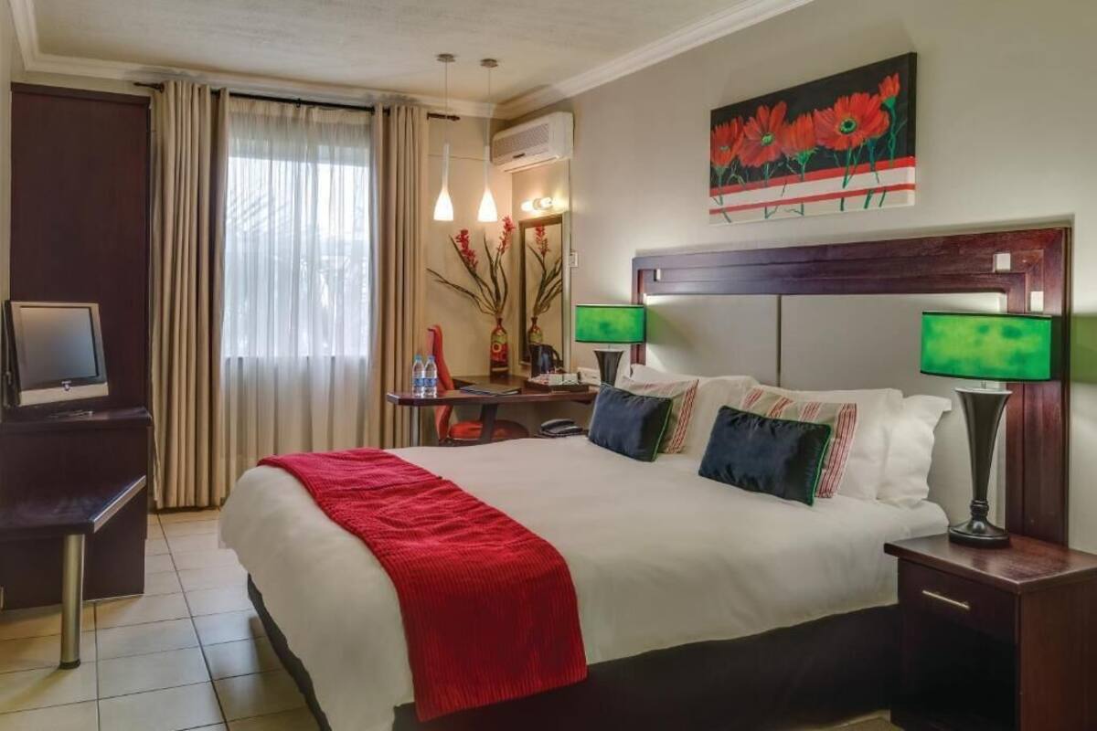 A hotel room with a bed, bedside lamps, a desk with a chair, a television, and a painting on the wall.