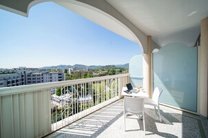 Uma varanda com uma cadeira branca, uma mesa com um laptop e um muro de vidro que oferece vista para edifícios e vegetação.