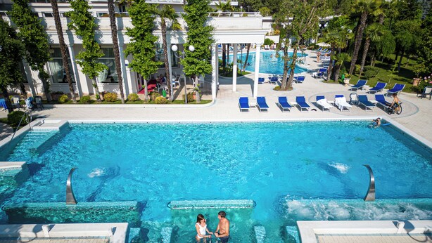 Une grande piscine extérieure avec deux personnes dans l’eau, entourée de chaises longues et de palmiers.