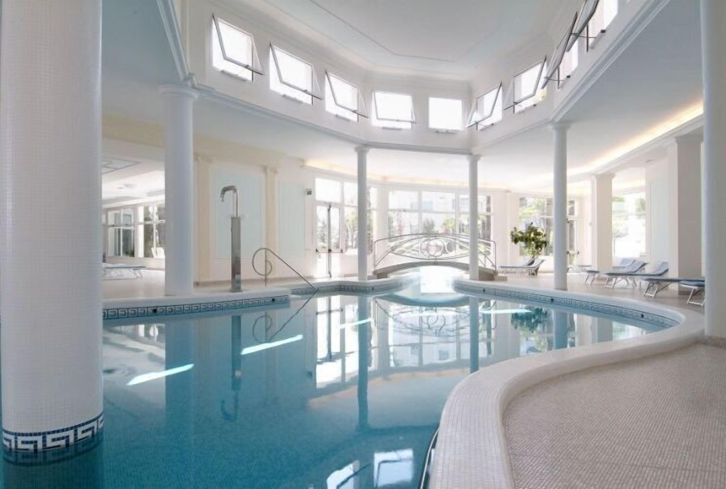 A spacious indoor pool with a curved design, surrounded by white columns and large windows.